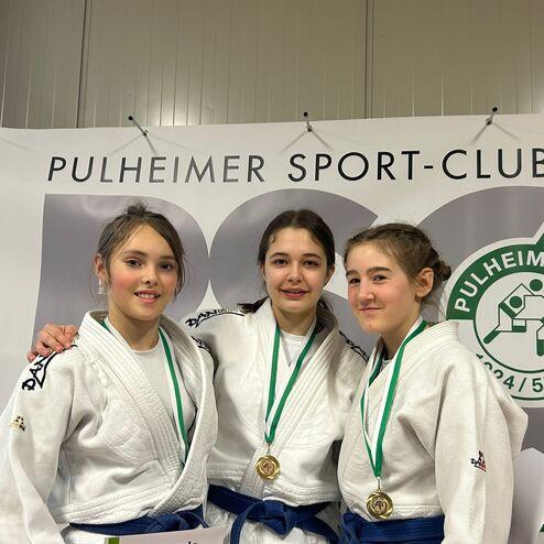 Drei Judokas in weißen Gi mit Medaillen posieren lächelnd vor einem Banner des Pulheimer Sport-Clubs.