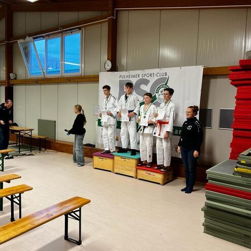 Vier Medaillenträger in Judogi stehen auf dem Siegerpodest, während Zuschauer Fotos machen und applaudieren.