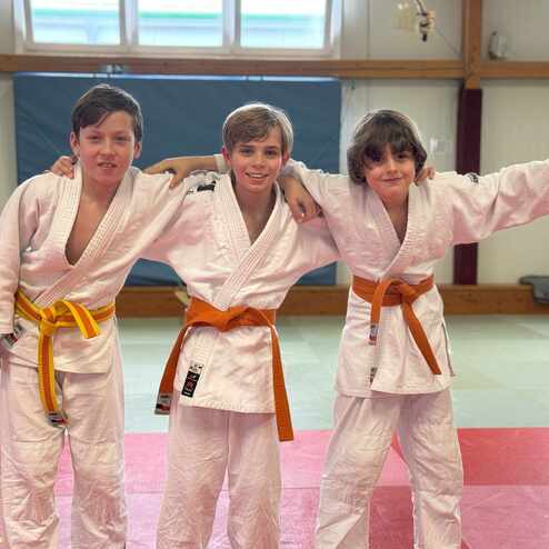 Drei Kinder in weißen Judoanzügen mit verschiedenen Gürtelfarben lächeln und posieren auf einer Judo-Matte.
