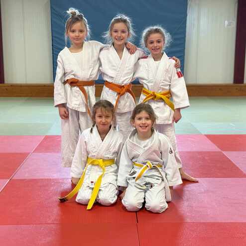 Fünf Kinder in Judogis stehen auf einer Judomatte; zwei tragen orange, drei gelbe Gürtel.