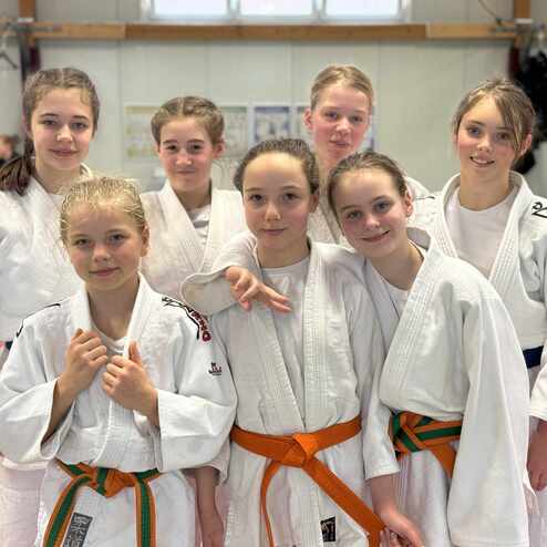Gruppe von sieben jungen Judokas in weißen Kimonos, lächelnd und posierend in einer Sporthalle.
