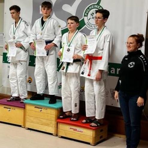 Vier Jugendliche in Judoanzügen stehen auf dem Siegertreppchen und halten Urkunden, während eine Trainerin danebensteht.