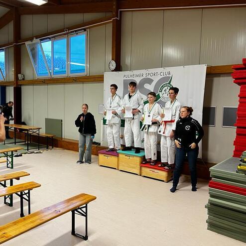 Siegerzeremonie im Judosport: Vier Medaillengewinner stehen auf dem Podium, während eine Frau spricht und Zuschauer filmen.