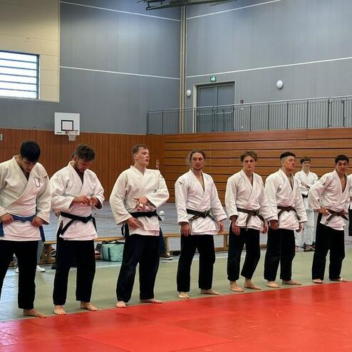 Sieben Judokas in wei&szlig;en Anz&uuml;gen mit schwarzen G&uuml;rteln stehen in einer Turnhalle auf einer roten Matte.