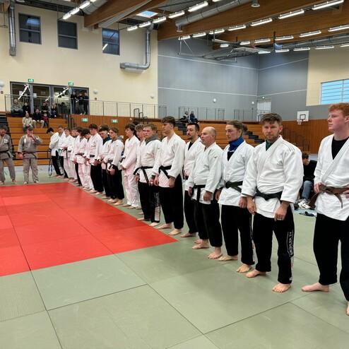 Gruppe von Judokas in wei&szlig;en und grauen Gi, auf einem Tatami, versammelt f&uuml;r eine Zeremonie in einer Sporthalle.