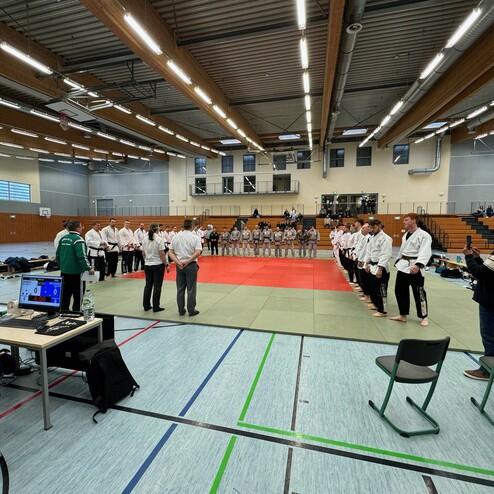Wettkampfhalle mit K&auml;mpfern in Judokleidung, die sich auf einer Matte versammeln, w&auml;hrend Zuschauer im Hintergrund sitzen.