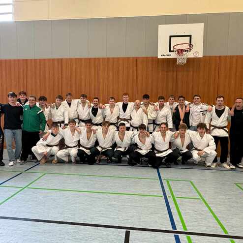 Gruppe von Judokas in wei&szlig;en und schwarzen Anz&uuml;gen posiert gemeinsam in einer Sporthalle. Basketballkorb im Hintergrund.