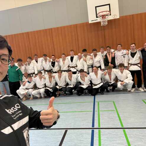 Selfie einer Person mit Brille vor einer Gruppe von Judoka in Trainingsanz&uuml;gen in einer Sporthalle.