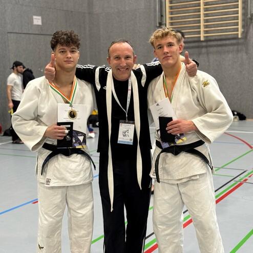 Zwei Judoka mit Medaillen stehen neben ihrem Trainer in einer Sporthalle. Alle drei l&auml;cheln in die Kamera.