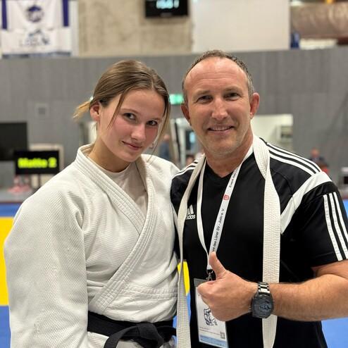 Junge Judoka in wei&szlig;em Gi posiert neben Trainer in schwarzem Shirt, beide l&auml;cheln vor einer Wettkampfumgebung.