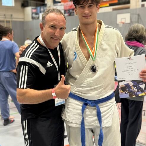 Junge Judo-K&auml;mpfer mit Medaille l&auml;chelt neben Trainer, h&auml;lt Urkunde in einer Sporthalle.