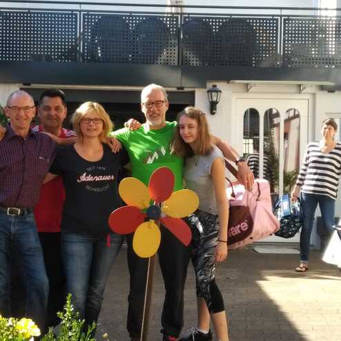 Gruppe von fünf Personen lächelt vor einem Gebäude, umgeben von Pflanzen und einem bunten Windrad.