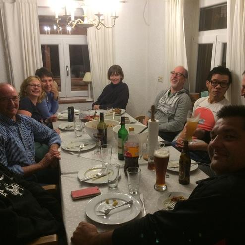 Gruppe von acht Personen sitzt an einem Tisch mit Tellern, Getränken und Essen in einem gut beleuchteten Raum.