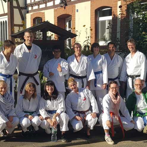 Gruppe von Karateka in weißen Anzügen und verschiedenen Gürtelfarben, posierend im Freien vor einem Gebäude.