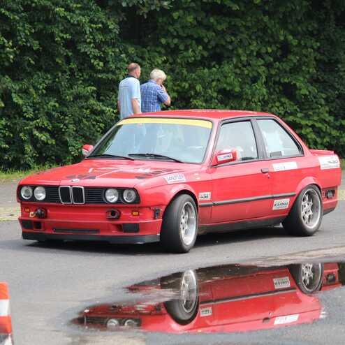 Roter BMW E30 fährt auf einer Strecke mit Wasserpfütze und zwei Personen im Hintergrund.