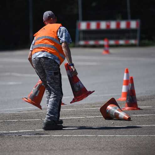 Zwei Personen in Warnwesten heben Verkehrshütchen auf einem Parkplatz mit verschiedenen Hutchen auf.