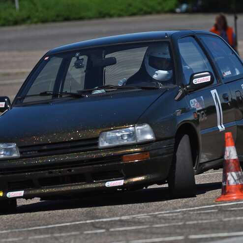 Schwarzes Auto mit Glitzer fährt um einen Pylonen auf einem Parkplatz bei sonnigem Wetter. Ein Fahrer trägt einen weißen Helm.