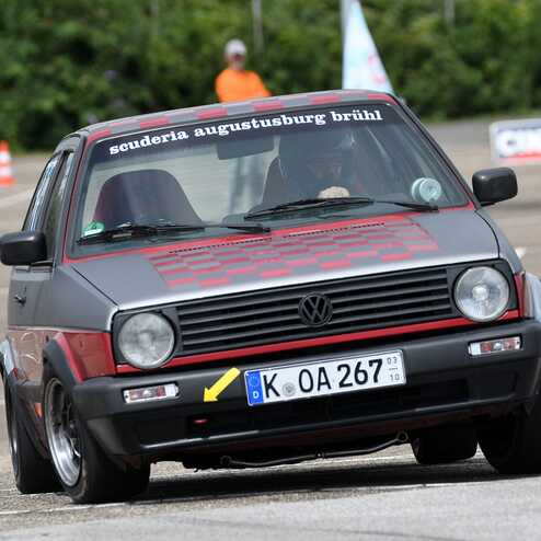 Volkswagen Golf im slalomartigen Fahrmanöver auf Parkplatz mit Pylonen und Zuschauer im Hintergrund.