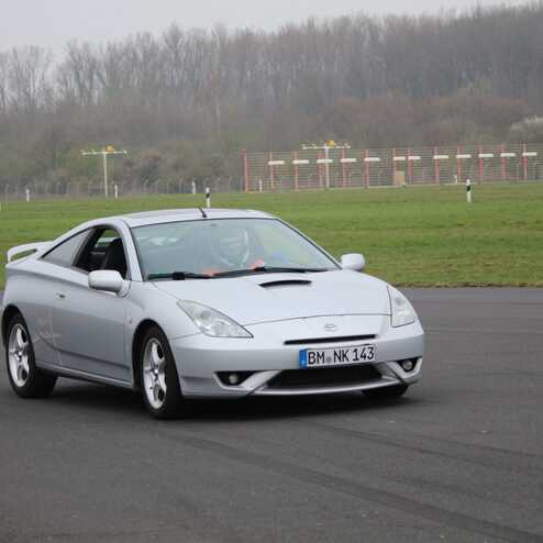 Silberner Sportwagen auf einer Rennstrecke, mit grünem Gras und Pylonen im Hintergrund. Fahrer im Cockpit sichtbar.