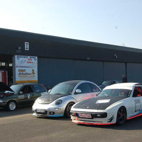 Drei Fahrzeuge stehen vor einer Werkstatt: ein grüner Kompaktwagen, ein silberner Beetle und ein getunter Sportwagen in Weiß.