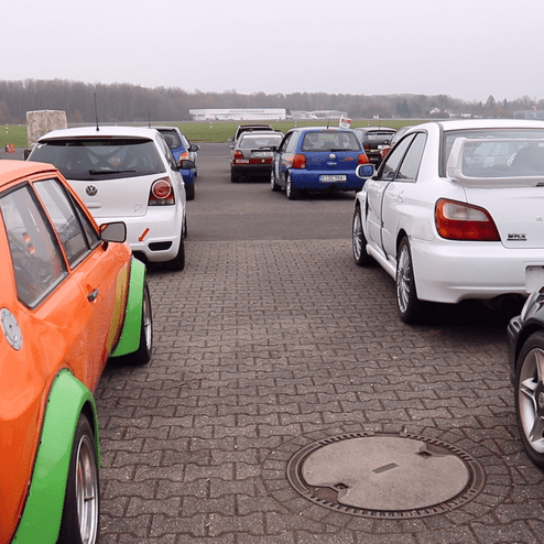 Bunte Autos parken auf einem Asphaltplatz; im Vordergrund ein orangefarbener Wagen mit grünen Details.