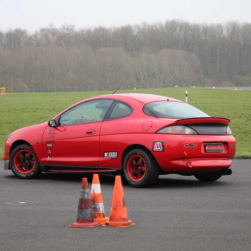 Roter Sportwagen fährt auf einem Übungsplatz, umgeben von Pylonen und einer grünen Wiese im Hintergrund.