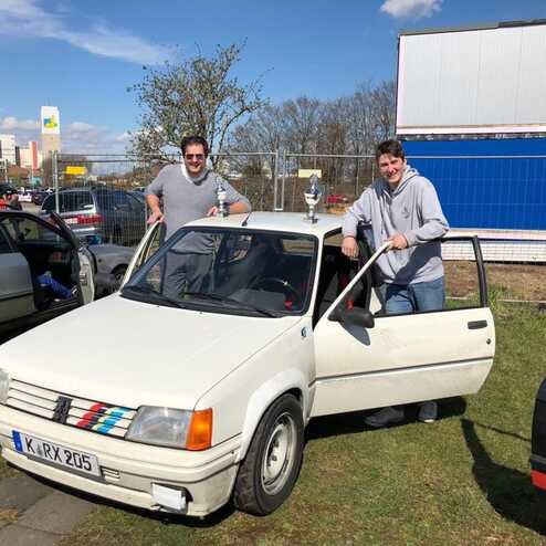 Zwei Männer stehen an einem weißen Auto mit Pokal, umgeben von weiteren Fahrzeugen und Baustellenhintergrund.