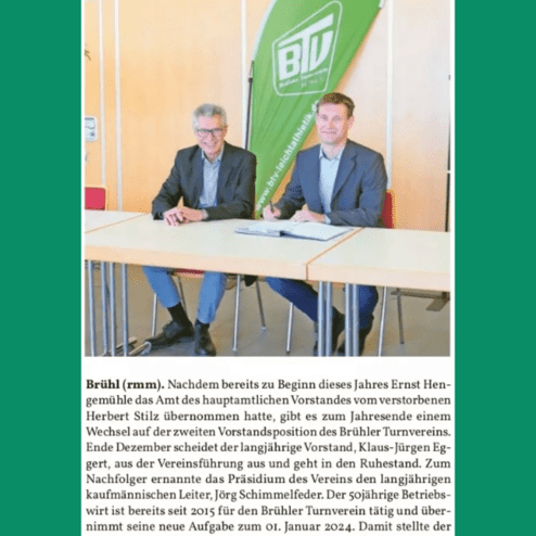 Zwei Männer in Anzügen sitzen an einem Tisch, mit einem grünen Banner des BTV im Hintergrund.