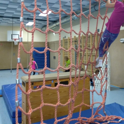 Mädchen hangelt an einem Seilnetz in einer Sporthalle, im Hintergrund Basketballkorb und Trainingsgeräte sichtbar.