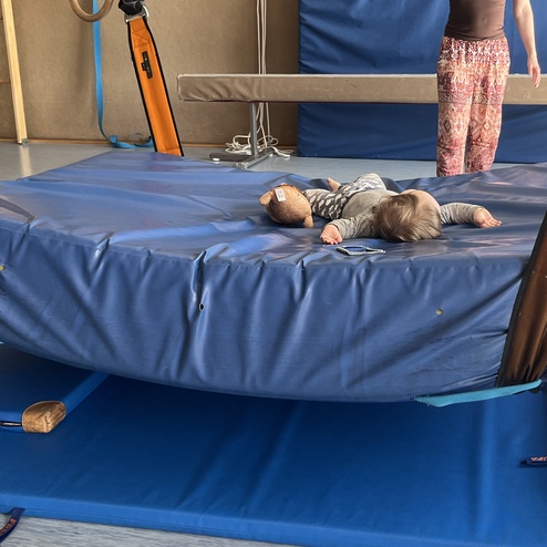 Zwei Kinder liegen auf einer blauen Turnmatte unter einem hängenden, gepolsterten Gerät, eine Erwachsene steht daneben.