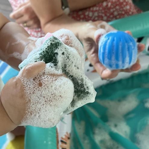 Kleines Kind hält einen schaumigen grünen Schwamm in der Hand, während eine erwachsene Hand einen blauen Ball präsentiert.