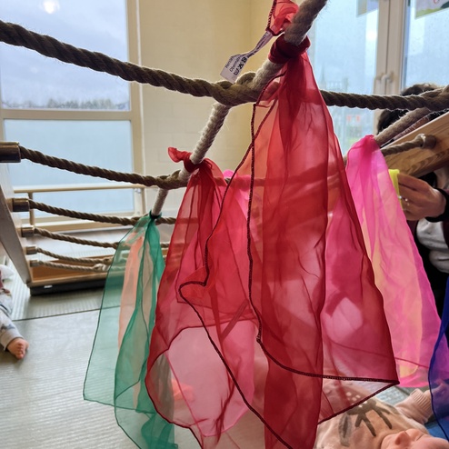 Bunte, transparente Tücher hängen an einem Seil innerhalb eines Raums, darunter ein spielendes Baby auf einer Matte.