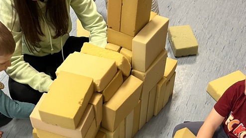 Kinder und eine Erwachsene bauen aus gelben Baukl&ouml;tzen eine Struktur auf einem blauen Boden.