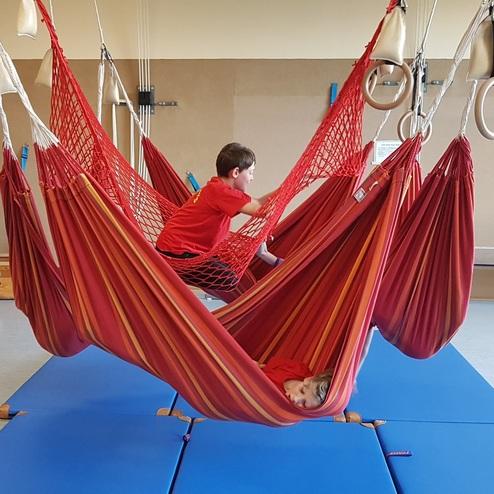 Zwei Kinder in bunten Hängematten spielen in einem Raum mit blauen Matten und Turngeräten.