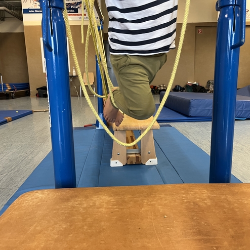 Kind schwingt an einem blauen Reck mit Seil, der Fuß steht auf einer Holzbank in einer Turnhalle.