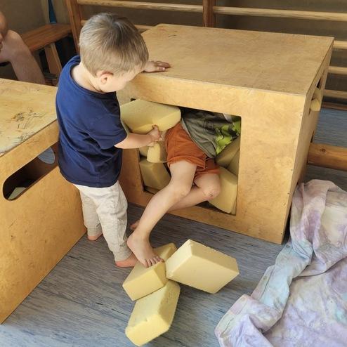 Ein kleiner Junge hilft einem anderen, der im Inneren einer Holzkiste mit Schaumstoffblöcken feststeckt.