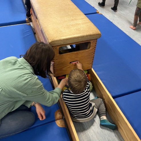 Ein Erwachsener hilft einem Kind, das in einem Holzspielzeug sitzt, während andere Kinder im Hintergrund spielen.