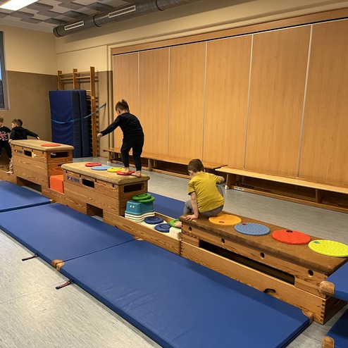 Kinder balancieren auf einem Holzgeräteparcours mit bunten Kreisen in einem Turnraum. Weiche Matten liegen darunter.