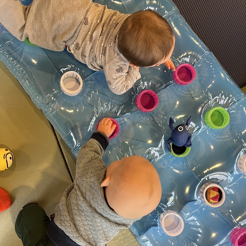 Zwei Babys spielen auf einer aufblasbaren Matte mit bunten Bechern und Spielzeug.