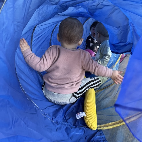 Zwei Kleinkinder sitzen in einem blauen Tunnelspielzeug, eines hält ein Plüschtier und schaut überrascht.