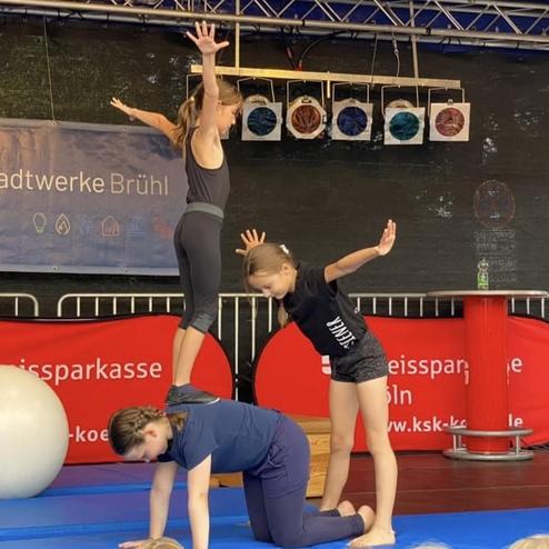 Drei Kinder führen eine akrobatische Übung auf einer blauen Matte vor einer Kulisse mit Sponsorenbanner aus.