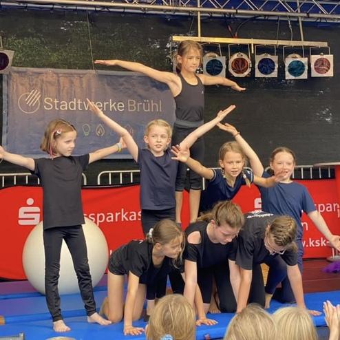 Acht Kinder in Sportkleidung führen eine Akrobatik-Formation auf einer blauen Matte bei einer Veranstaltung auf.