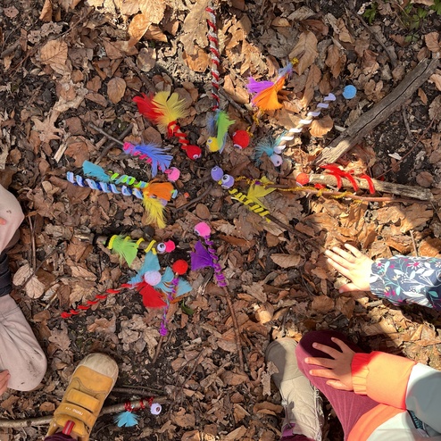 Bunte Holzfiguren und Materialien liegen auf Laub, umgeben von Kinderhänden, die aktiv beim Gestalten einbezogen sind.