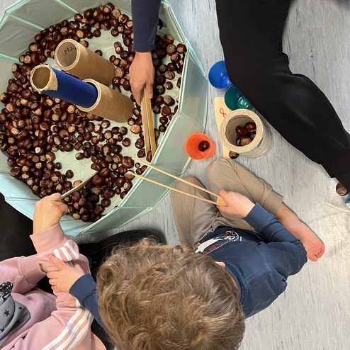 Zwei Kinder spielen mit Kastanien und Rohren in einem großen, kreisförmigen Behälter auf einem blauen Boden.