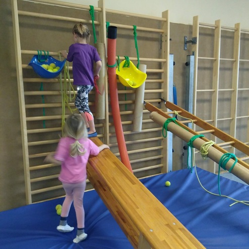 Zwei Kinder bewegen sich im Gymnastikraum auf einer Holzbank und klettern an einer Wand hoch, um gelbe Bälle zu erreichen.