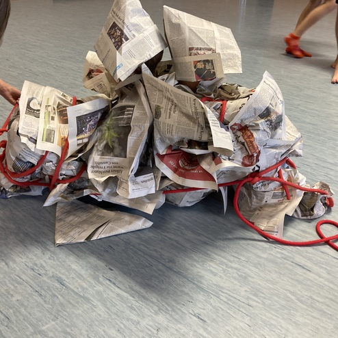 Kind bindet Zeitungspapier mit rotem Seil in Form eines Hügels auf einem blauen Boden. Weitere Kinder sind im Hintergrund sichtbar.