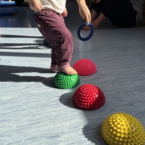 Kind steht barfuß auf einem grünen, spitzen Spielball und greift nach einem blauen Ring. Rote und gelbe Bälle liegen daneben.