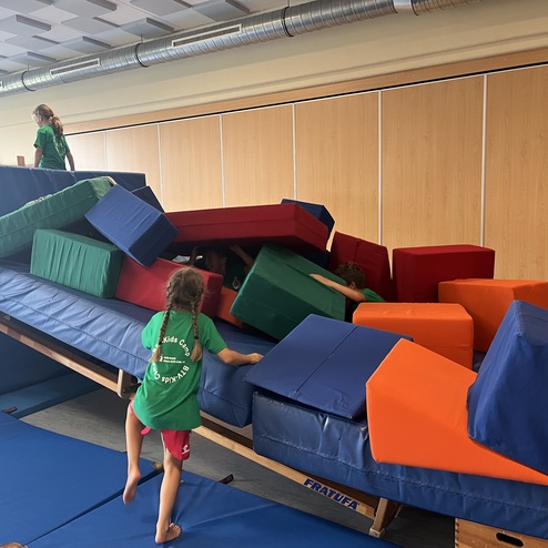 Kinder spielen auf einem bunten Hindernisparcours aus Matten und Gymnastikblöcken in einer Turnhalle.
