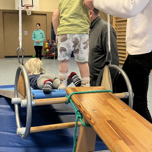 Kinder spielen in einer Turnhalle, einige sind auf einer Schaukel, andere von Erwachsenen betreut. Bunte Bälle im Hintergrund.