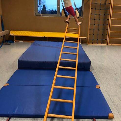 Mädchen schwingt an Ringen über einer Holzleiter, die auf blauen Turnmatten platziert ist. Sporthalle mit Fenster im Hintergrund.